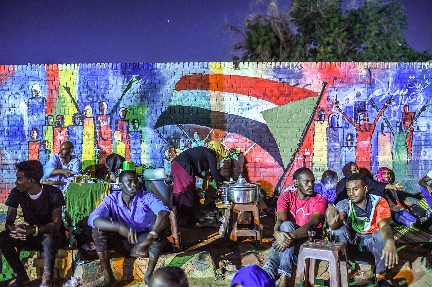 سودانيون يجلسون أمام جدارية تجسد الاحتجاجات الشعبية في الخرطوم، 24 نيسان/أبريل 2019. (أ ف ب)