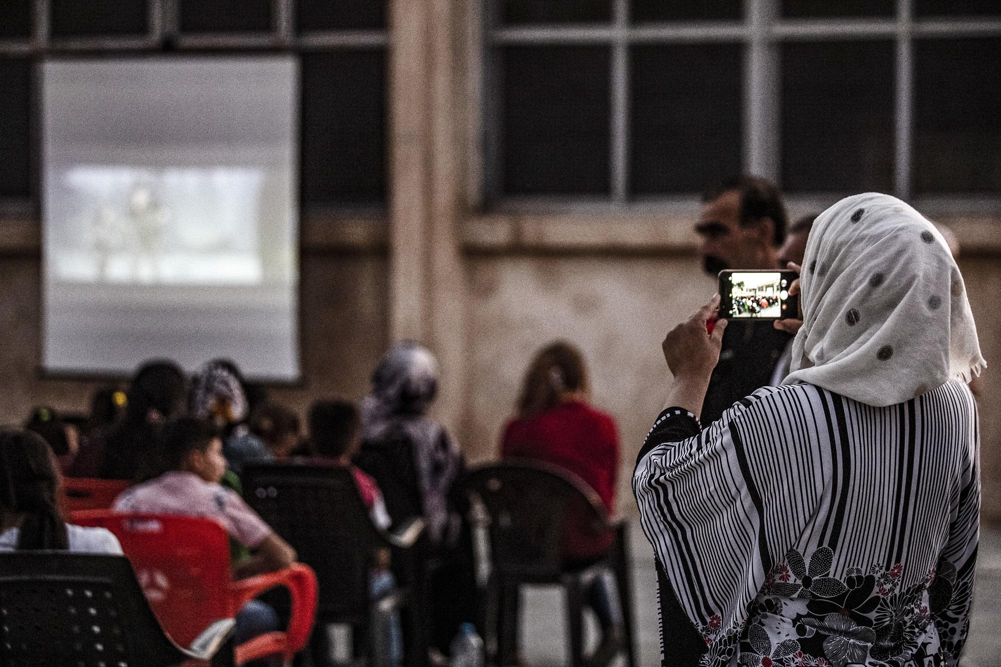 سيدة تستخدم هاتفًا خلويًا لتصوير فيديو لأطفال يتابعون عرضا للسينما المتنقلة في باحة مدرسة في الحسكة، شمال شرق سوريا. 28 يوليو 2019. (أ ف ب)