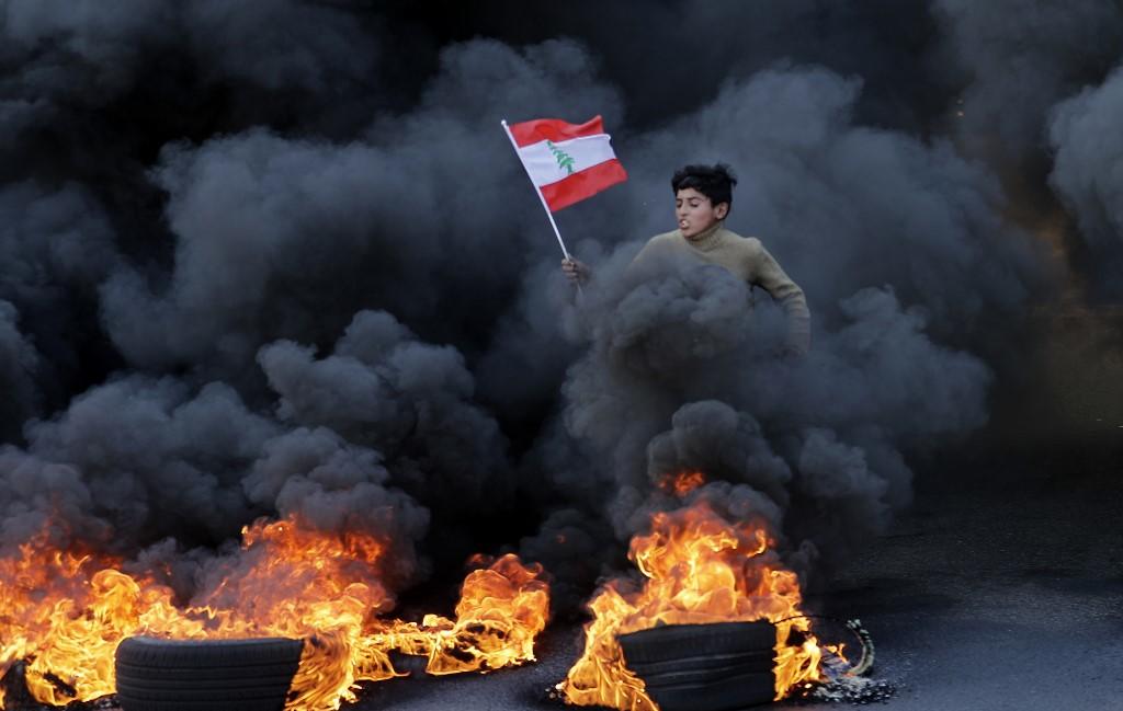 شاب لبناني يركض حاملاً العلم الوطني بينما يتصاعد الدخان من الإطارات المحترقة أثناء مظاهرة في منطقة جل الديب على المشارف الشمالية للعاصمة اللبنانية بيروت، 14 يناير/كانون الثاني 2020. (جوزيف عيد/ أ ف ب)