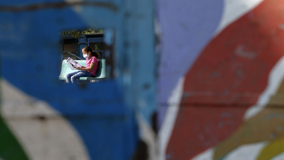 فتاة فلسطينية ترتدي كمامة للوقاية من فيروس كورونا المستجد، تظهر من خلال فتحة في باب مدرسة بينما تقرأ كتابها في مخيم النصيرات وسط قطاع غزة. 16/11/2020, (محمد عابد / أ ف ب)