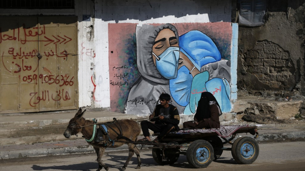 فلسطيني وزوجته يركبان عربة يجرها حمار أمام لوحة جدارية تظهر أطباء يرتدون كمامات واقية؛ بسبب جائحة فيروس كورونا في مخيم النصيرات وسط قطاع غزة. 16/11/2020. (محمد عابد / أ ف ب)