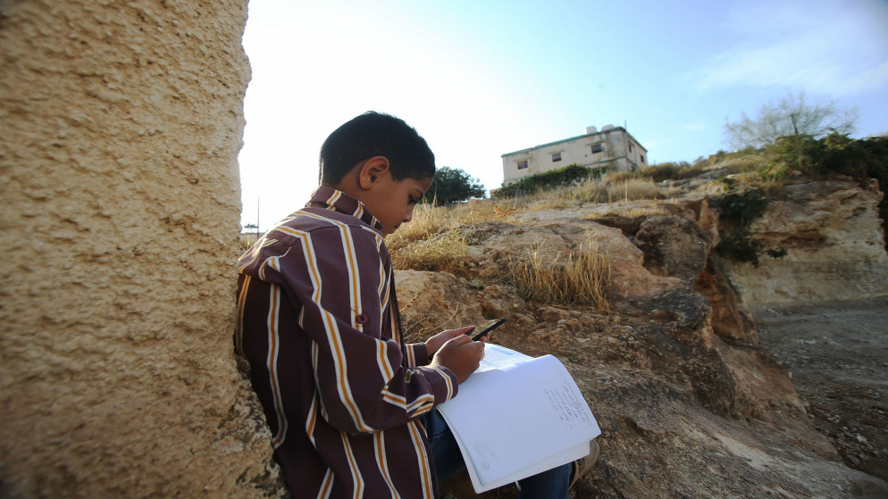 طالب يتابع دروسه عبر هاتف محمول، يجلس بقرب منزل أهله في محافظة المفرق بحثا عن تغطية شبكة إنترنت. (صلاح ملكاوي / المملكة)