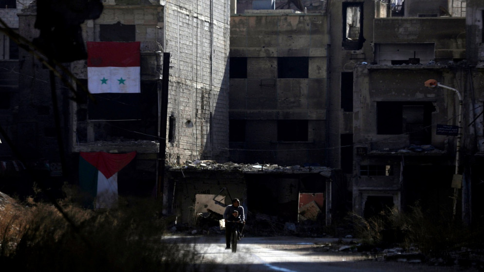 رجل يركب دراجة مع طفل أمام مبنى مدمر مزين بالأعلام السورية والفلسطينية في مخيم اليرموك للاجئين الفلسطينيين في الضواحي الجنوبية لدمشق، سوريا، 2 كانون الأول/ ديسمبر 2020. (رويترز)
