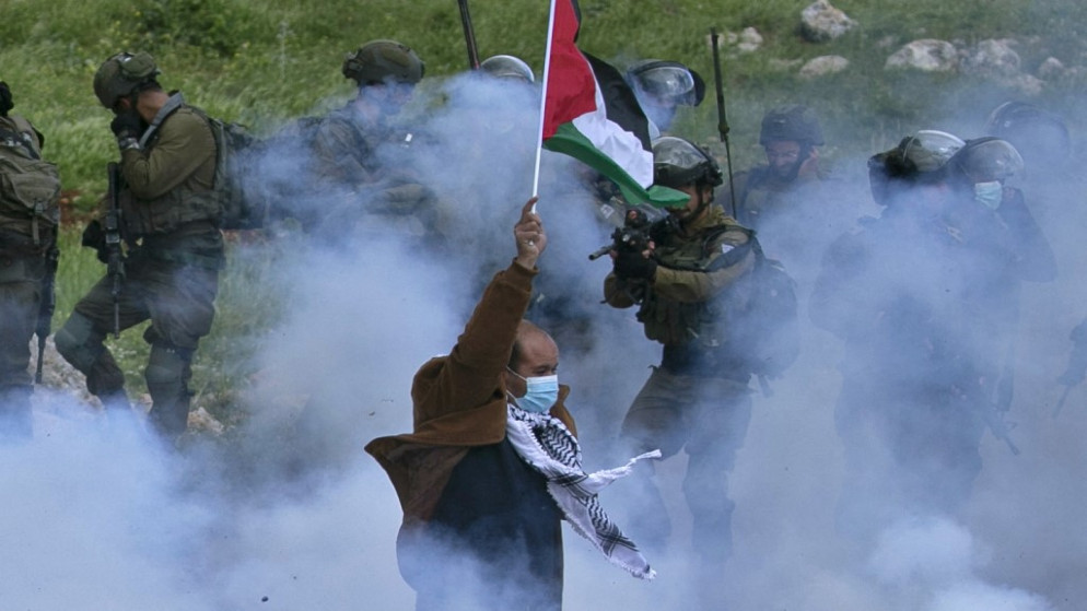 فلسطيني يرفع العلم بينما يصوب جندي إسرائيلي بندقيته عليه وسط دخان الغاز المسيل للدموع في الضفة الغربية المحتلة . 2 نيسان /أبريل 2021.(أ ف ب)