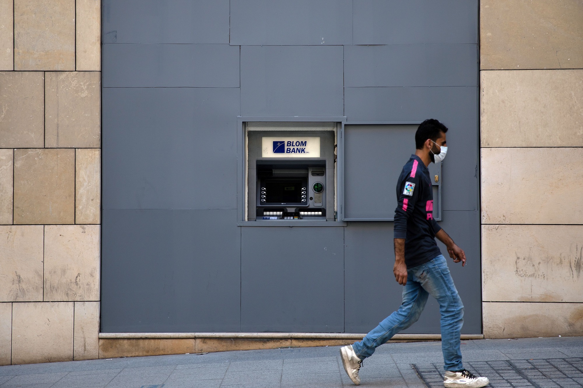 رجل يرتدي كمامة للوجه للوقاية من فيروس كورونا المستجد يسير أمام ماكينة صراف آلي في بيروت. (رويترز)