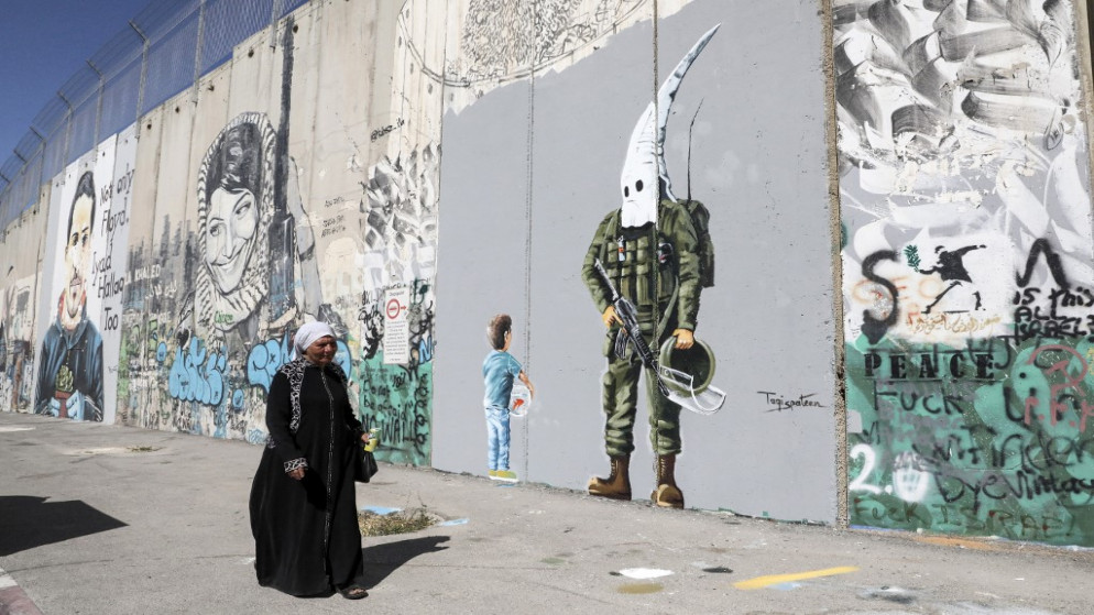 فلسطينية تمر أمام لوحة جدارية مرسومة على جدار الفصل العنصري تصور صبيًا يحمل حوض سمك بينما ينظر إليه جندي الاحتلال الإسرائيلي الذي يرتدي زياً عسكريا.الضفة الغربية المحتلة. 30 مايو/أيار 2021.(أ ف ب)