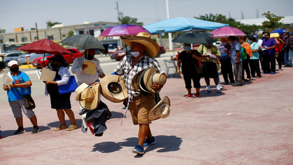 رجل يبيع قبعات بينما يصطف الناس لتلقي لقاح فيروس كورونا، خلال برنامج تطعيم شامل للأشخاص الذين تزيد أعمارهم على 50 عامًا في ملعب بيسبول في سيوداد خواريز بالمكسيك، 22/6/2021. (رويترز / خوسيه لويس جونزاليس)
