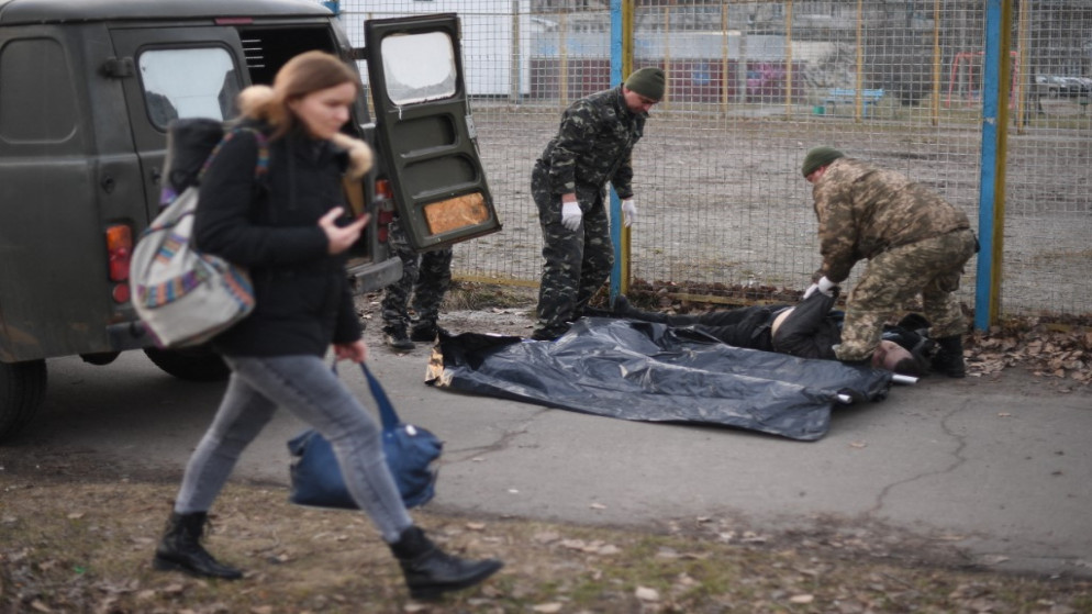 امرأة تمشي بينما يستعد الجنود الأوكرانيون لالتقاط جثة رجل أوكراني أصيب برصاصة عندما مرت عربة مصفحة روسية أمامه، على رصيف في شمال كييف .25 فبراير 2022. (أ ف ب)