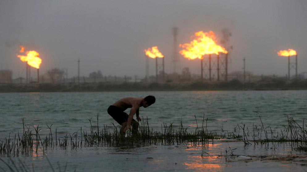 رجل يقف في نهر شط العرب وألسنة اللهب تتصاعد من أنابيب مصفاة النفط في الخلفية في البصرة، العراق، 23 تموز/ يوليو 2020. (رويترز)