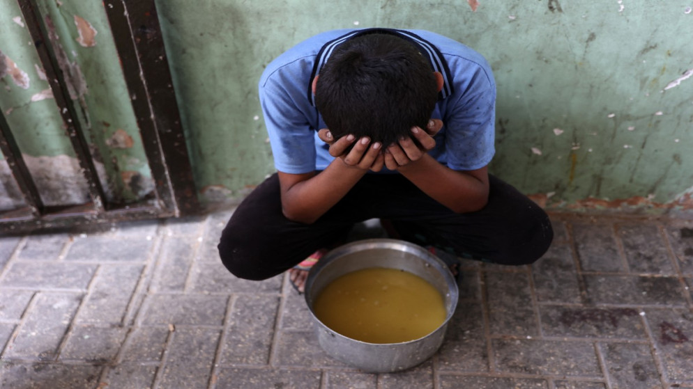 طفل فلسطيني نازح يجلس بجانب قدر من حساء العدس تسلّمه في نقطة توزيع طعام بمدينة غزة. 25 تموز 2025. (أ ف ب)