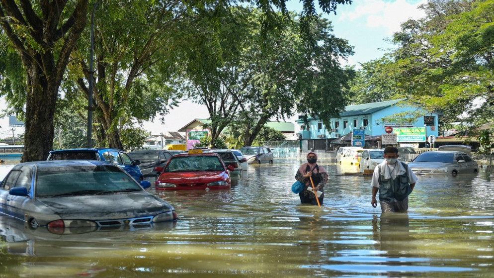 أشخاص يمشون بجوار سيارات غارقة جزئيًا في مياه الفيضانات في شاه علم، سيلانجور، 21 ديسمبر 2021. (أ ف ب)