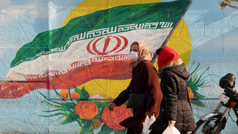 إيرانيات يسرن في شارع رئيسي في العاصمة طهران وخلفها جدار رسم عليه العلم الوطني لإيران. 28/12/2021. (أ ف ب)